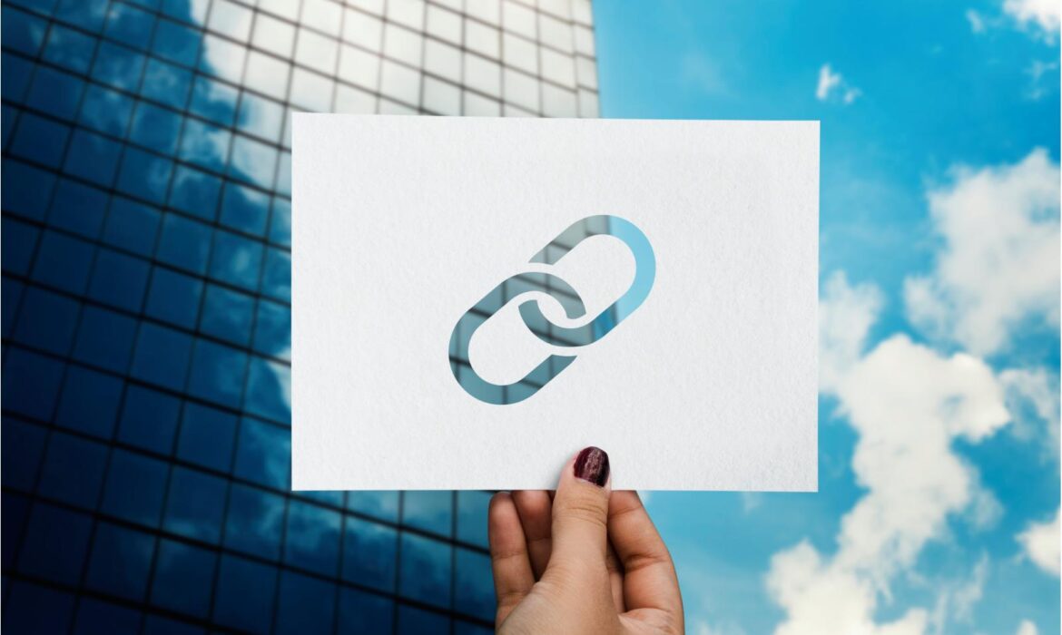 A hand holds a paper with a chain link symbol cutout, against a backdrop of a modern glass building and blue sky with clouds, conveying connectivity.