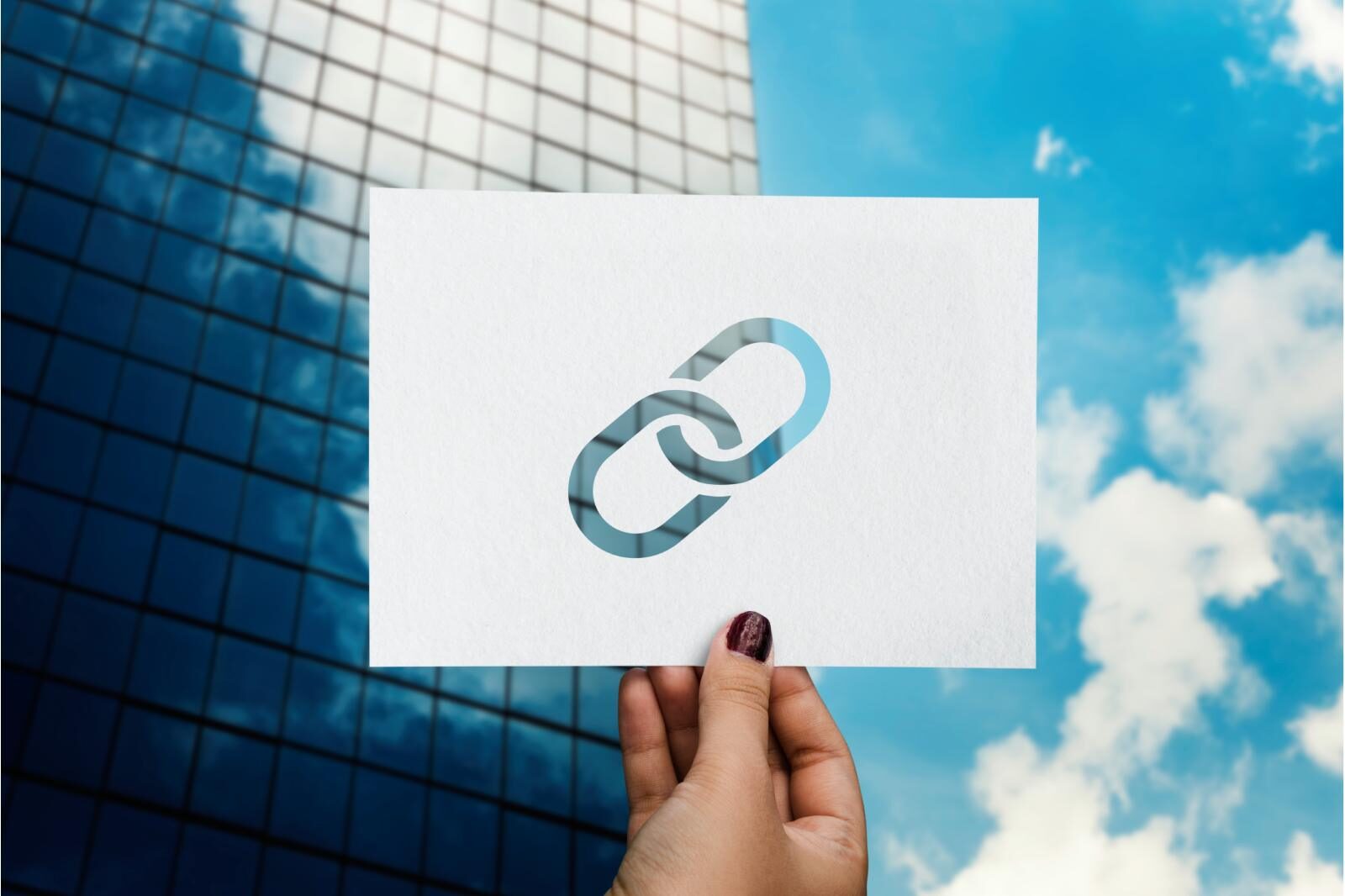 A hand holds a paper with a chain link symbol cutout, against a backdrop of a modern glass building and blue sky with clouds, conveying connectivity.